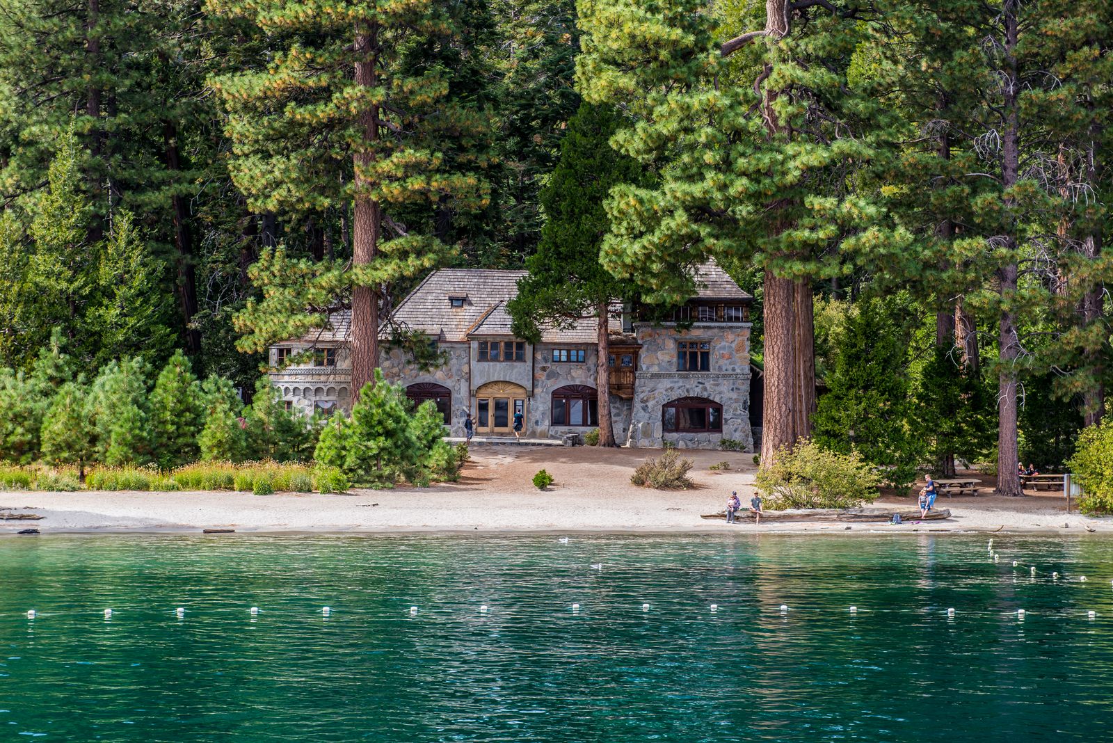 Stone lakeside house beneath tall pines on clear blue water